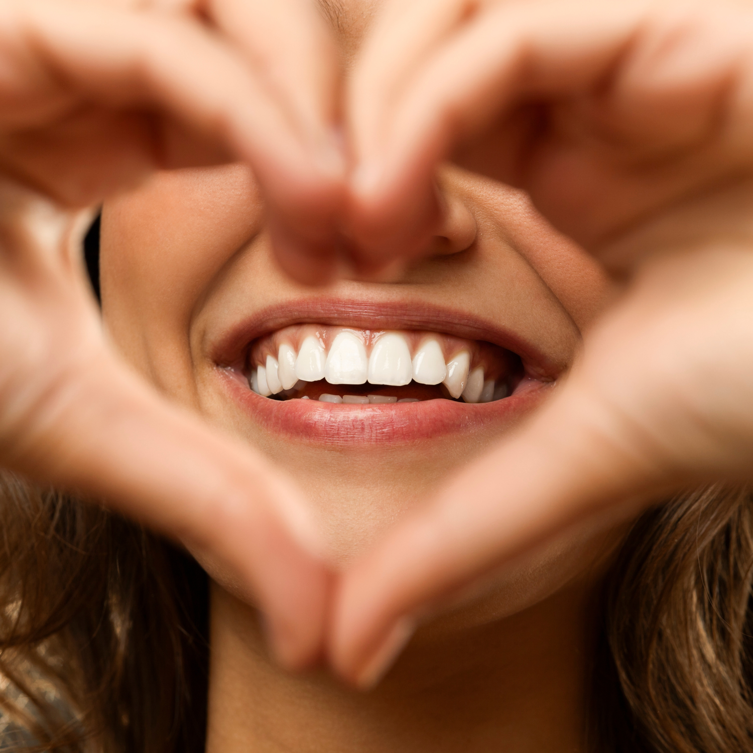 Close-up of a white, healthy smile with hands forming a heart shape, symbolizing brighter teeth and the OrellaUv Brighter Smile collection