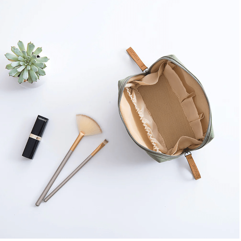 Open green makeup bag with beige interior, lip gloss, brush, and small plant on a white background